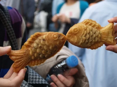 Kaksi henkilöä pitää ylhäällä taiyaki-leivoksia niin, että näyttää siltä kuin kalat suutelisivat